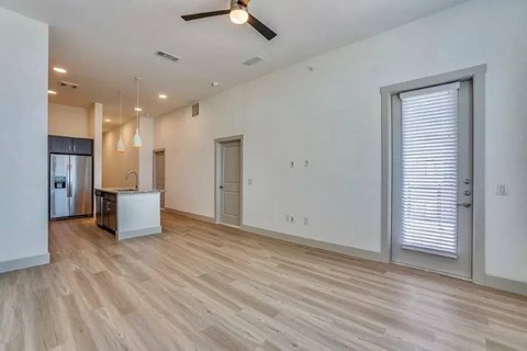 A bright open-concept living and kitchen area here at Valor at the Realm with light wood flooring, white walls, a modern island with pendant lights, stainless steel refrigerator, and a glass door with blinds.