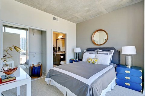 A bedroom here at Victory Place, including a nighstand, modern lamp, a mirror above the bed, with a sliding  closet in the wall. The space is encompassed by bright natural light