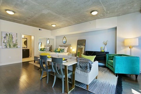 A modern open studio here at Victory Place with dark wood flooring, a dining table, cozy seating, green accent furniture, and a sleeping area with decorative bedding and nearby bathroom access.