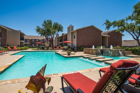 A resort-style outdoor pool here at Wildflower featuring clear blue water, red cushioned lounge chairs, shaded umbrellas, and a stone water feature, surrounded by landscaped greenery and apartment buildings, creating a relaxing and inviting community space for residents to enjoy warm days outdoors.