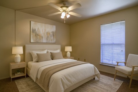 A calm, modern bedroom here at Wildflower featuring a neutral upholstered bed with crisp white bedding, textured accent pillows, and a soft throw blanket. Coordinated nightstands with matching lamps, a ceiling fan with lighting, plush carpeting, and a large window with blinds create a comfortable, restful retreat filled with natural light.