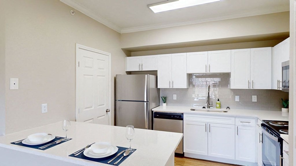 Modern kitchen here at Landing at Mansfield Apartment Homes with white cabinets, stainless steel appliances, and a glossy backsplash. An island with two place settings adds a clean, inviting feel.