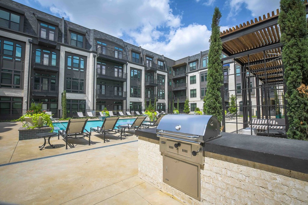 Modern The Crosby at The Brickyard Apartment Homes courtyard with a pool, lounge chairs, barbecue grill, and pergola. The setting is serene and sunny, with lush greenery.