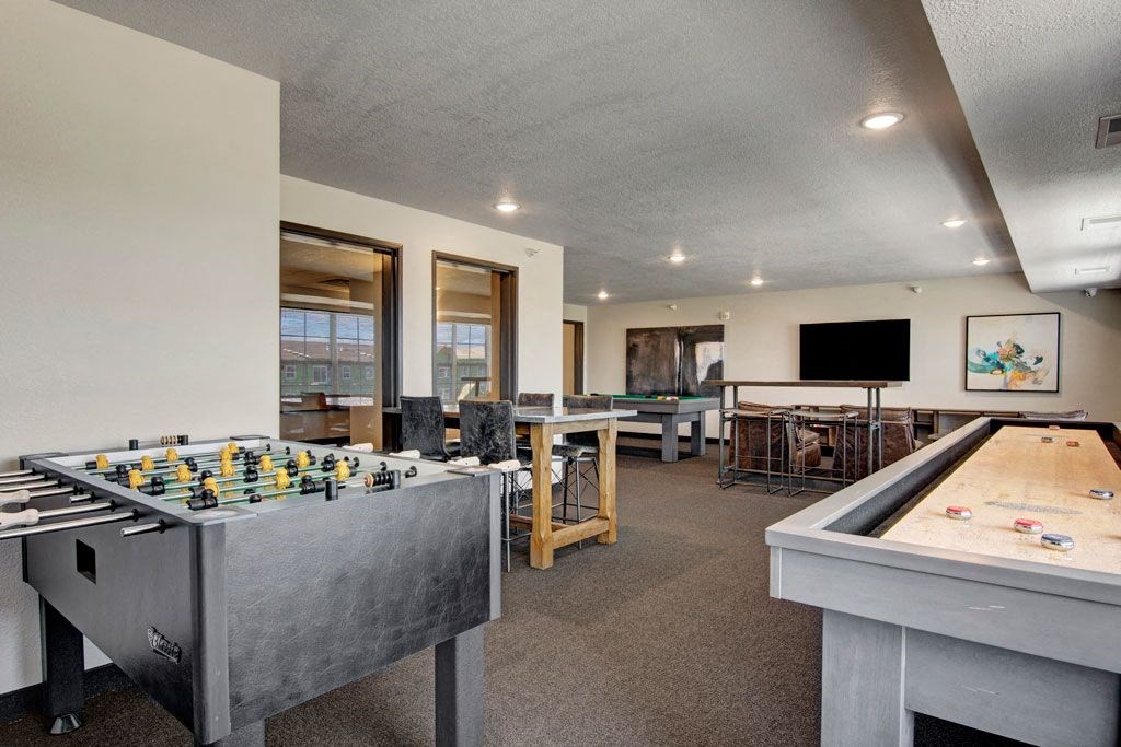Game room here at The Club at Heritage Apartment Homes with a foosball table in the foreground, shuffleboard on the right, and a TV on the wall. Bright, spacious, and neatly arranged.