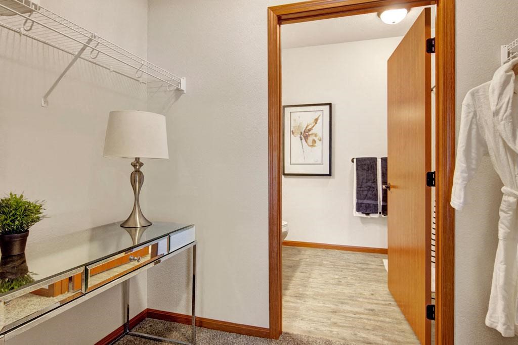 Modern dressing area here at The Club at Heritage Apartment Homes with mirrored vanity, decorative lamp, and potted plant on plush carpet. Open door reveals bathroom with abstract art. Cozy atmosphere.