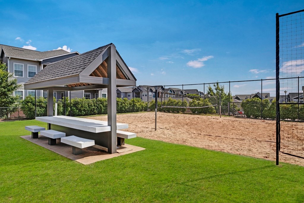 Picnic area here at Lincoln at Central Park Apartment Homes with a covered table and benches on green grass, adjacent to a sandy volleyball court, set against residential buildings under a clear blue sky.