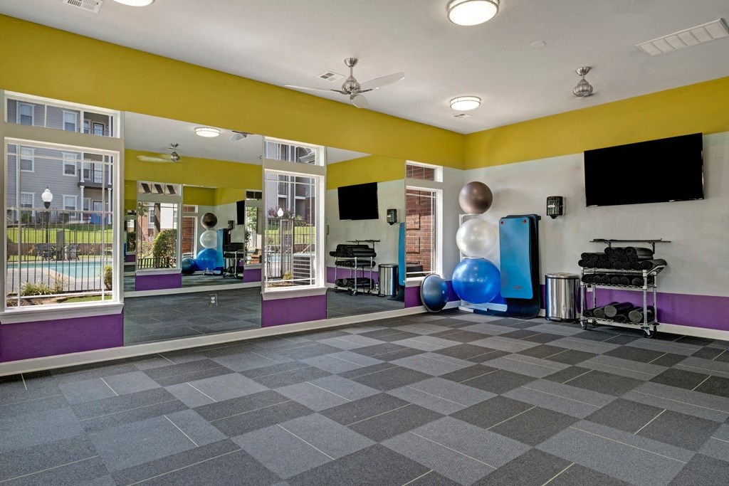 Bright, spacious fitness room here at Lincoln at Central Park Apartment Homes with mirrored wall, yoga mats, exercise balls, and weights rack. Large windows show outdoor pool area. Calm, inviting atmosphere.