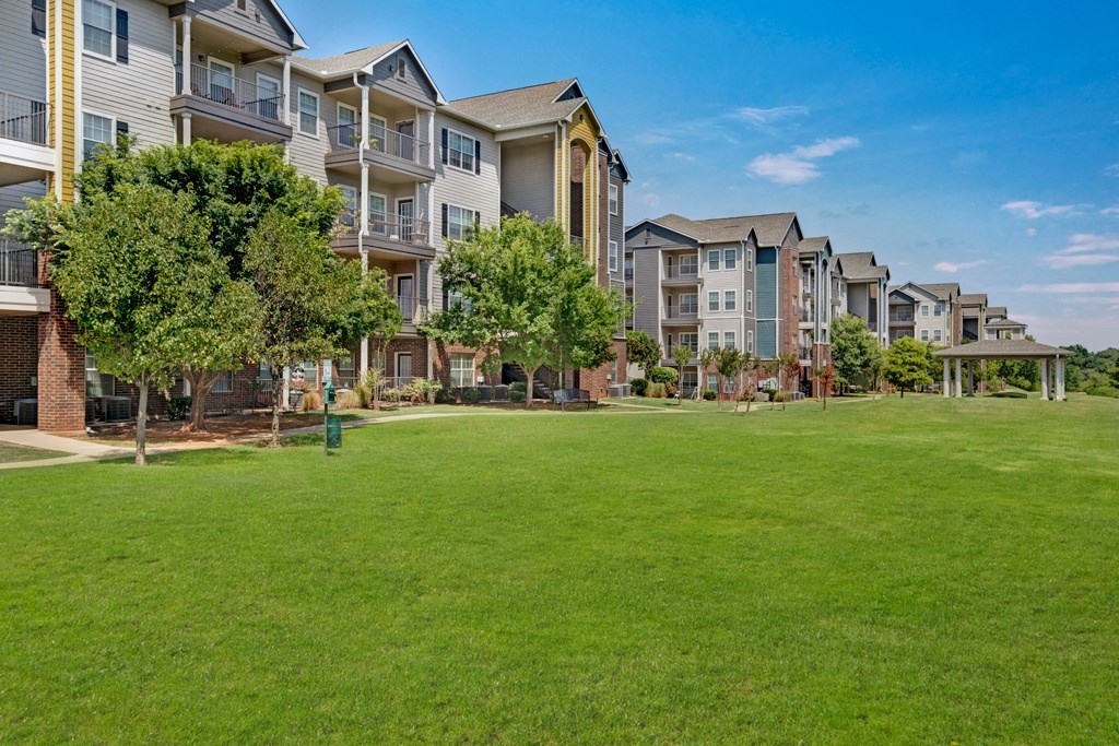 A sunny day view of modern Lincoln at Central Park Apartment Homes lined with trees, adjacent to a large green lawn. The scene conveys a serene, suburban atmosphere.