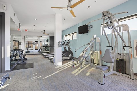 Bright gym here at The Uptown Apartment Homes with blue walls featuring treadmills, stationary bikes, and weight machines. Ceiling fans provide ventilation, creating an inviting, energetic atmosphere.