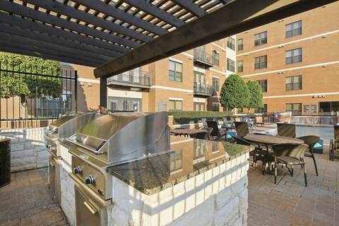 Outdoor patio here at The Uptown Apartment Homes with modern stainless steel grills under a pergola, surrounded by tables and chairs. Apartment buildings and a tree in the background.