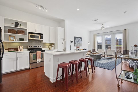 Modern open-plan kitchen here at The Uptown Apartment Homes and living area with hardwood floors. White cabinets, stainless steel appliances, bar stools, and cozy lounge with large windows.