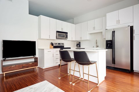 Modern kitchen here at The Uptown Apartment Homes with white cabinets, stainless steel appliances, and an island with black chairs. Hardwood floors add warmth and a cozy feel.
