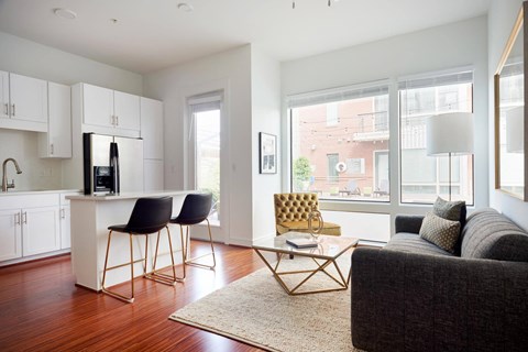 Modern living room here at The Uptown Apartment Homes with large windows, white walls, and hardwood floors. Features a gray sofa, yellow chair, sleek coffee table, and kitchen bar. Bright, airy ambiance.