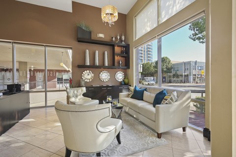 A modern, elegant living room here at The Uptown Apartment Homes with beige walls and large windows. Features white furniture, decorative shelves, a chandelier, and a tranquil, cozy atmosphere.