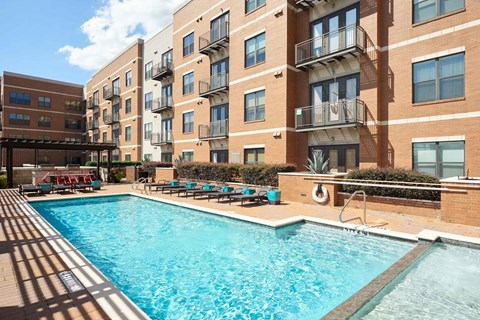 The Uptown Apartment Homes building with multiple balconies overlooks a shimmering outdoor pool surrounded by lounge chairs, under a partly cloudy blue sky.