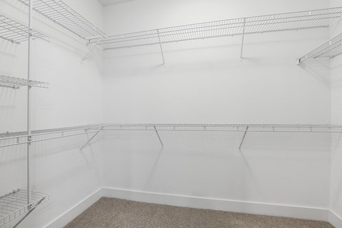 Empty white walk-in closet here at The Uptown Apartment Homes with multiple wire shelves along the walls, beige carpeted floor. The space feels clean, bright, and spacious.