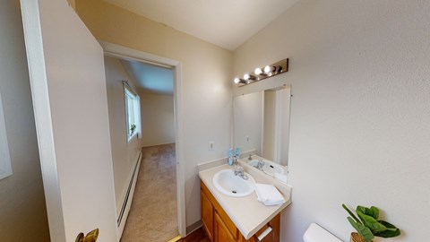 Bright bathroom here at the Village at Calais Apartments with beige walls, a wood cabinet sink, and a large mirror. A corridor with carpet and a window leads to another room.