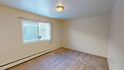 Spacious, empty bedroom here at the Village at Calais Apartments with beige carpet and walls, large window with vertical blinds, and a small plant on the sill. Overhead light fixture provides a cozy feel.