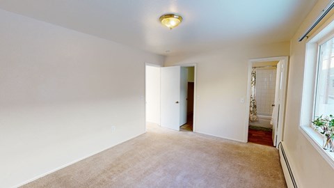 Empty bedroom here at the Village at Calais Apartments with beige carpet, cream walls, and a ceiling light. Open doors reveal a closet and a bathroom with a white tiled shower curtain. A large window adds brightness.