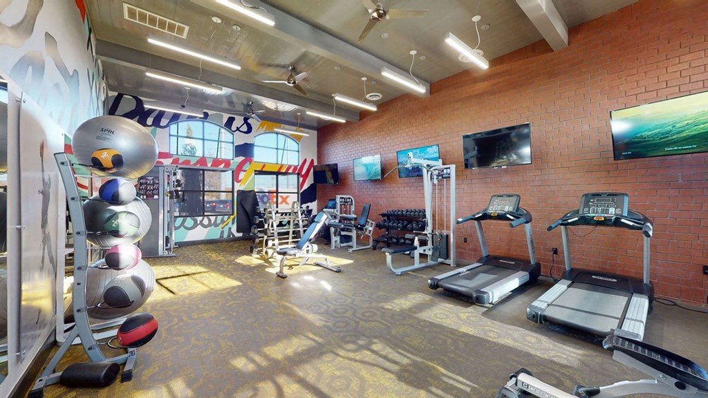Modern gym here at 71 at Tulsa Hills Apartment Homes with colorful mural walls features treadmills, weight machines, dumbbells, and exercise balls. Large windows and bright lighting create an energetic atmosphere.