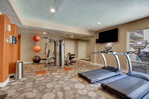 A fitness room here at Bridges with treadmills, a cable weight machine, wall mirrors, mounted TV, patterned carpet, and orange accent walls with exercise balls and strength equipment.