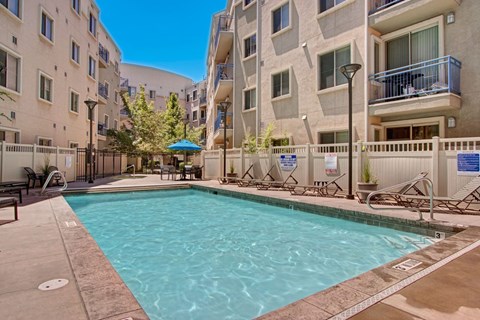 A pool courtyard here at Bridges with a rectangular pool, beige apartment buildings, blue balcony railings, tan deck, and lounge chairs, creating a bright, inviting outdoor amenity space.