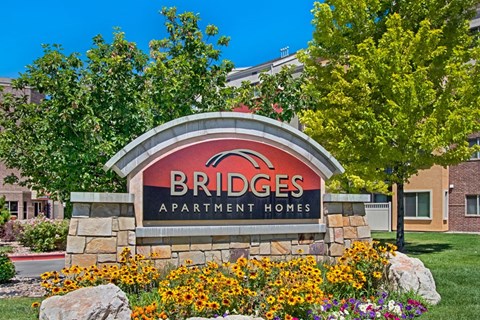 A community monument sign here at Bridges with a stone base and curved red-and-black Bridges Apartment Homes panel, surrounded by bright yellow flowers, green trees, and landscaped greenery under a clear blue sky.
