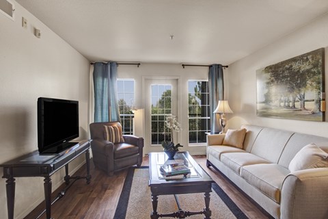 A living room here at Legends at River Oaks with a light neutral sofa, brown leather accent chair, blue curtains, glass coffee table, dark TV console, and bright balcony doors bringing in natural light.