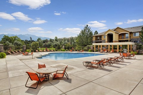 A pool area here at Legends at River Oaks with a large blue pool, orange lounge chairs and tables, wide concrete deck, pergola seating, and mountain views under a bright sky.