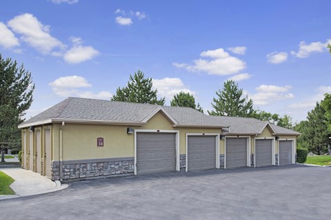 A detached garage building here at Legends at River Oaks with multiple gray garage doors, tan and stone exterior walls, dark asphalt drive, and green trees behind under a bright blue sky.
