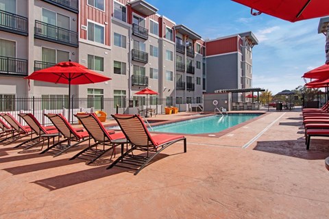 A sunny outdoor pool area here at Ritz Classic with a large swimming pool, red lounge chairs and umbrellas, spacious deck seating, and modern apartment buildings surrounding the courtyard for a lively, resort-style atmosphere.