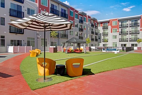 A vibrant courtyard lawn here at Ritz Classic with green turf, yellow lounge chairs, striped umbrellas, shaded cabana seating, and modern apartment buildings surrounding the space, creating a bright, social outdoor gathering area.