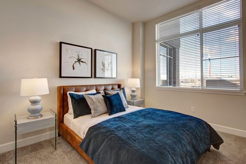 A bright bedroom here at Ritz Classic with a warm brown upholstered bed frame, blue bedding and accent pillows, matching nightstands with lamps, soft carpet, and a large window with blinds bringing in natural light and city views.