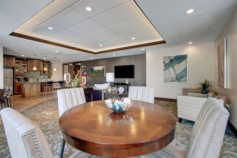 A spacious resident lounge here at Velo on the Boulevard with a round wood dining table, upholstered chairs, comfortable seating, wall-mounted TV, and an open community kitchen area, creating a warm, social space for gathering and relaxing.