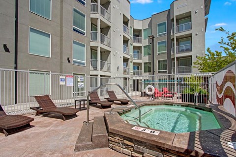 A private outdoor spa here at Velo on the Boulevard with a heated hot tub, metal handrails, surrounding lounge chairs, and a secured courtyard setting beside modern apartment buildings, creating a relaxing amenity space.