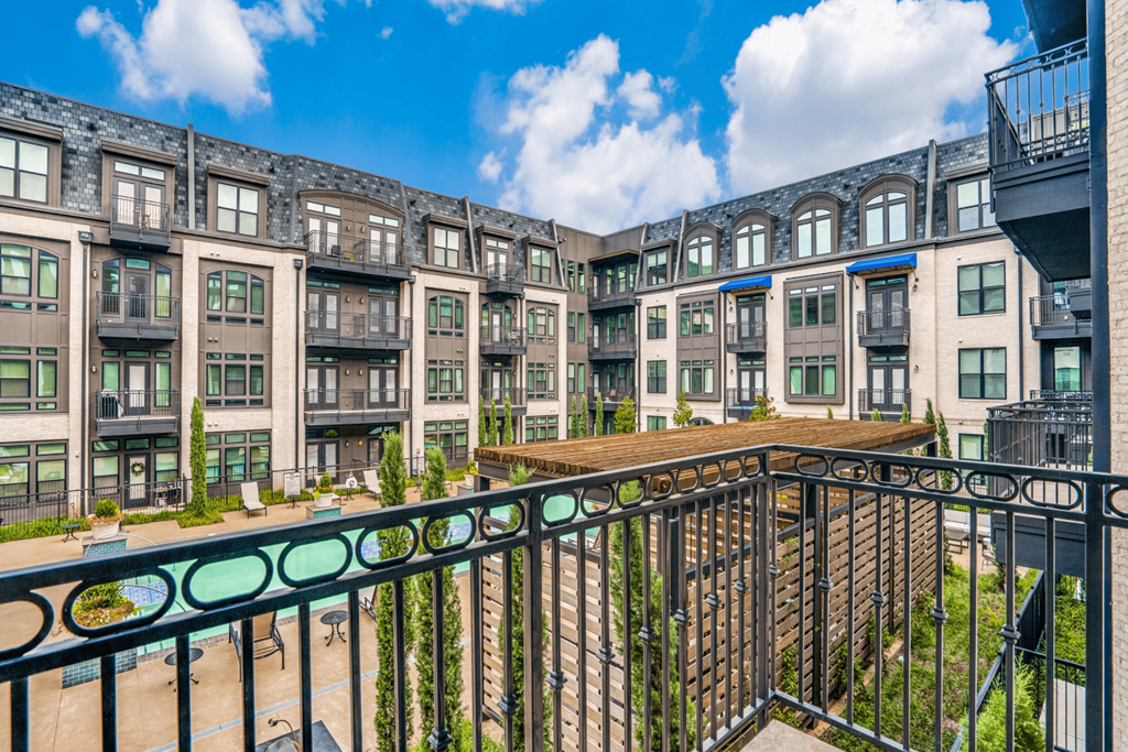 View from a balcony here at The Crosby at the Brickyard Apartment Homes overlooking a modern apartment complex. Features include large windows, small balconies, a pool, and a clear blue sky with fluffy clouds.