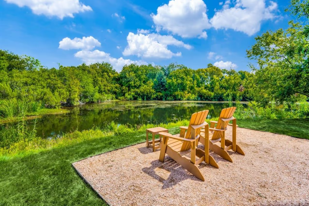 Two wooden chairs on a gravel patio overlook a serene pond surrounded by lush greenery here at The Crosby at The Brickyard Apartment Homes. The sky is bright blue with fluffy clouds. Peaceful scene.