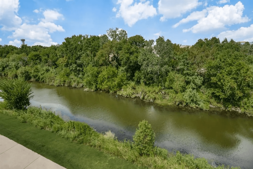 Scenic view of a calm river here at The Crosby at The Brickyard Apartment Homes, bordered by lush green trees under a bright blue sky with fluffy clouds. Serene and peaceful atmosphere.
