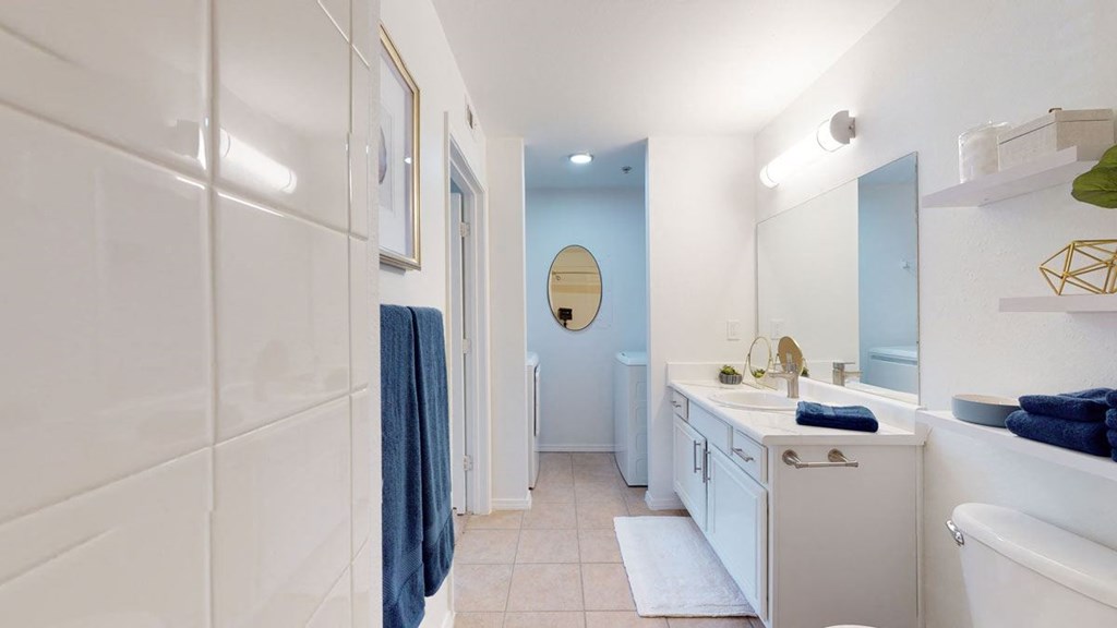 Modern bathroom here at Villas at Preston Lakes Apartment Homes with white walls and tiled floor. Features a double vanity with mirror, blue towels, and decorative items. Bright, clean, and inviting.