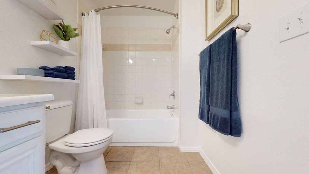 A clean, modern bathroom here at Villas at Preston Lakes Apartment Homes with a white bathtub and shower curtain, beige tiles, and blue towels on a rack and shelves. Neutral, tidy, and inviting space.