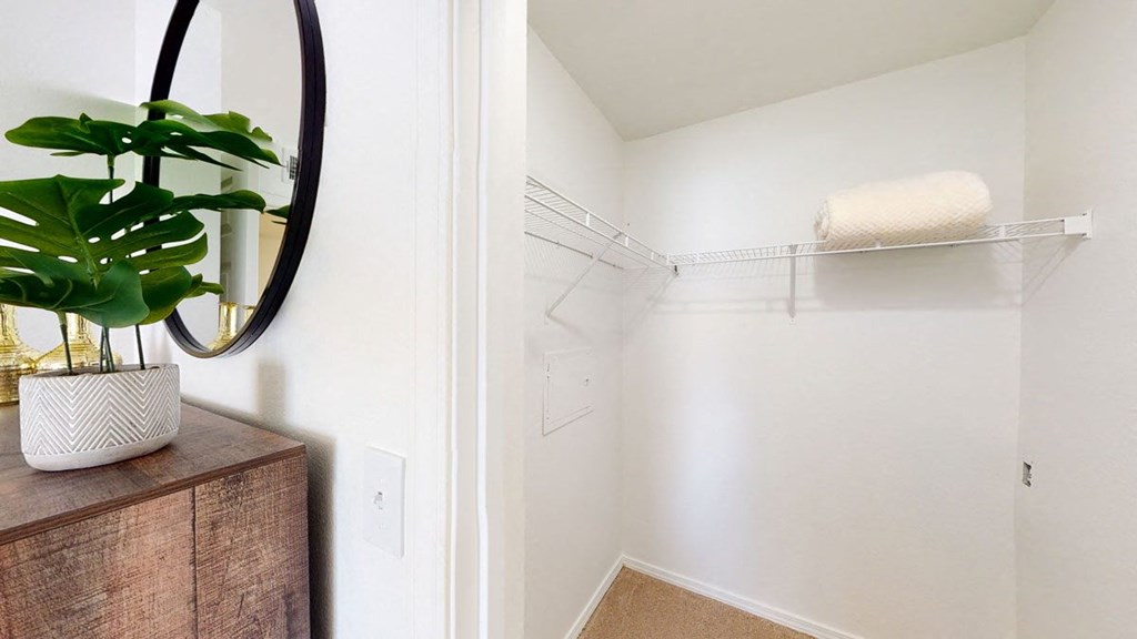Minimalist white closet here at Villas at Preston Lakes Apartment Homes with wire shelf holding a rolled cream blanket. Foreground shows a mirror and potted plant on a wooden cabinet, creating a clean, serene look.