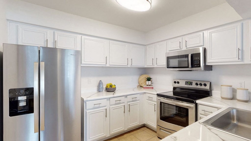 Modern kitchen here at Villas at Preston Lakes Apartment Homes with white cabinets, stainless steel appliances, and a light tile floor. A fruit bowl and plant add a cozy touch to the countertop.