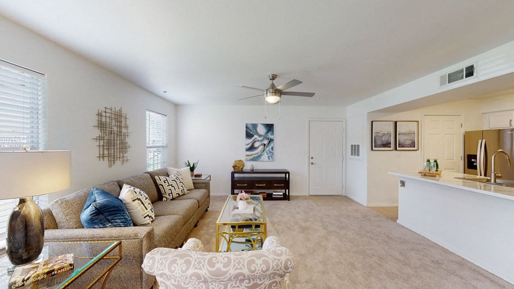 Modern living room here at Villas at Preston Lakes Apartment Homes with beige carpet and walls, featuring a comfy sofa with patterned pillows, a glass coffee table, abstract art, and a ceiling fan.