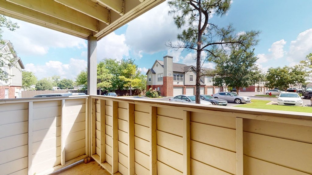 View from a small balcony here at Villas at Preston Lakes Apartment Homes with wooden railing, overlooking a parking lot and townhouse complex. Blue sky and fluffy clouds create a serene atmosphere.