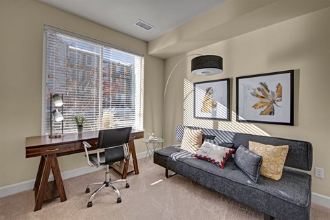 A room with a grey couch, a desk with a chair, and two framed pictures on the wall.