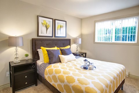 A cozy bedroom here at Colonial Square featuring a neatly made bed with a patterned yellow and white comforter, layered accent pillows, and a padded headboard, flanked by matching nightstands with modern lamps, complemented by framed wall art, soft carpeted flooring, and a window with vertical blinds providing natural light.