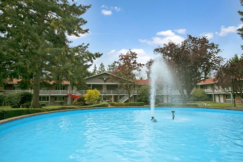 A serene fountain courtyard here at Colonial Square showcasing a large circular pool with a tall central water feature, surrounded by mature evergreen and deciduous trees, manicured hedges, and open green lawn areas, with two-story apartment buildings and exterior walkways framing the peaceful, park-like community setting.