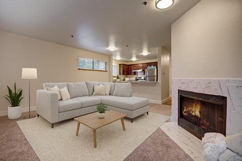 A modern living room here at Forest Greens featuring beige carpet, soft neutral walls, and an open layout that flows toward the kitchen. The space is staged with a light sectional sofa, neutral pillows, a wooden coffee table, a floor lamp, and greenery, while a tiled fireplace adds a cozy focal point.