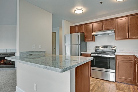 A spacious open kitchen here at Forest Greens featuring rich wood cabinets, light stone-style countertops, and a full appliance setup with a refrigerator, range, and hood. A wide breakfast bar overlooks the adjacent living area, while wood-look flooring and ceiling lights create a warm and functional cooking and gathering space.