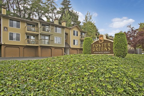 A welcoming exterior view here at Forest Greens showcasing the community monument sign set among manicured greenery, with multi-story apartment buildings in warm neutral tones behind it. The scene features private balconies, garage access below the residences, mature trees, and a landscaped setting that creates a peaceful residential atmosphere.